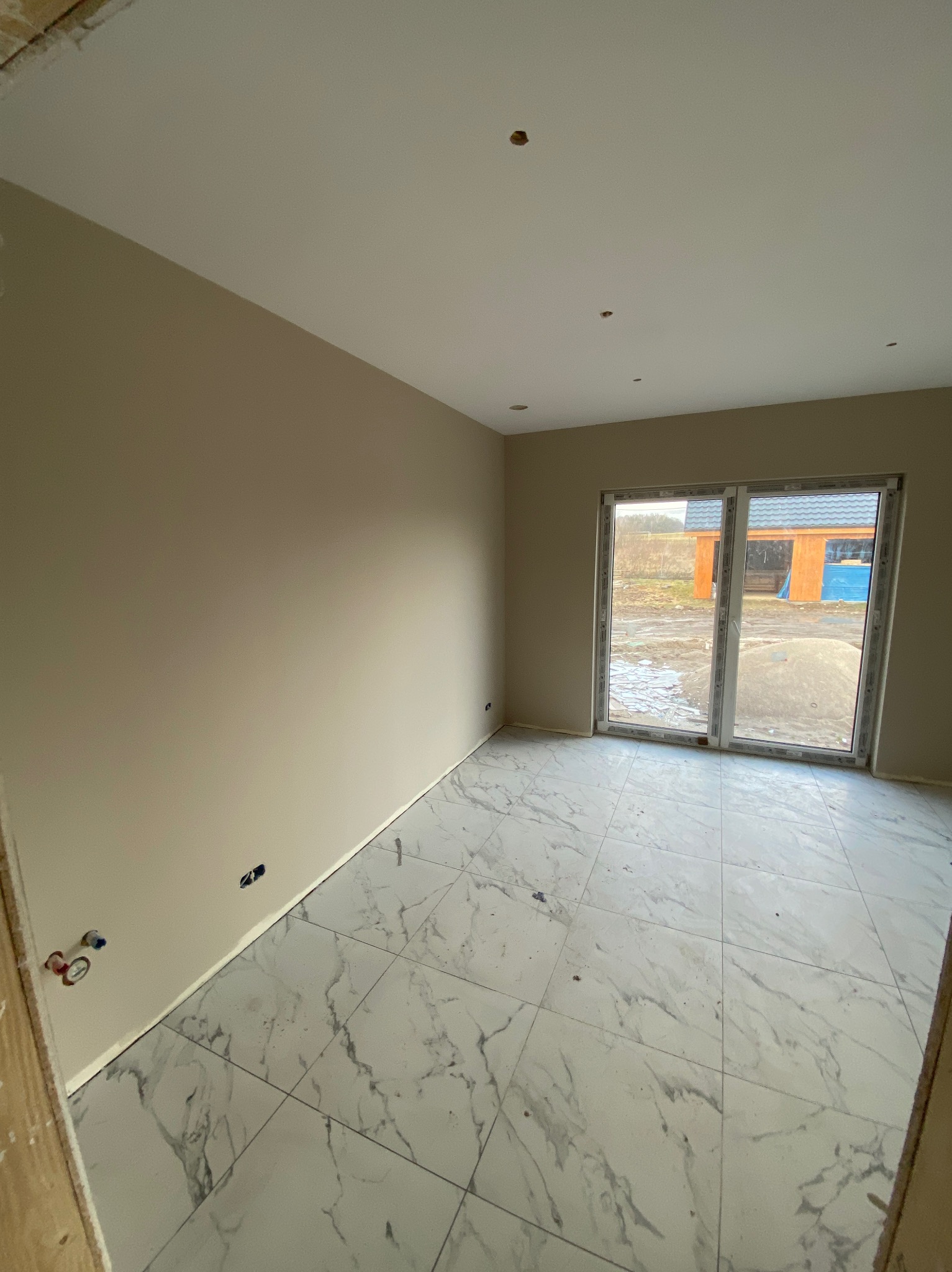 Jasne wnętrze po remoncie: beżowe ściany, białe marmurowe płytki podłogowe, duże okno z widokiem na zewnątrz. Widoczne gniazdka elektryczne i instalacja wodna.