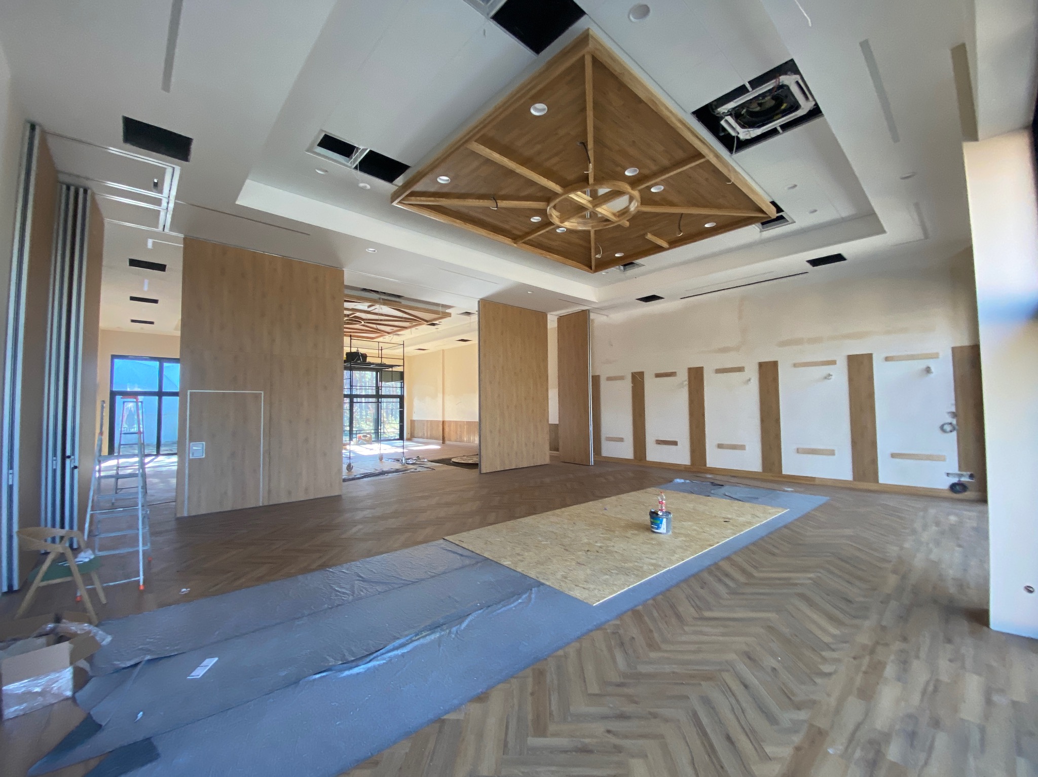 Wnętrze w trakcie prac wykończeniowych: parkiet w jodełkę zabezpieczony folią, ściany z panelami, drewniany sufit kasetonowy, drabina. Widoczne elementy instalacji.