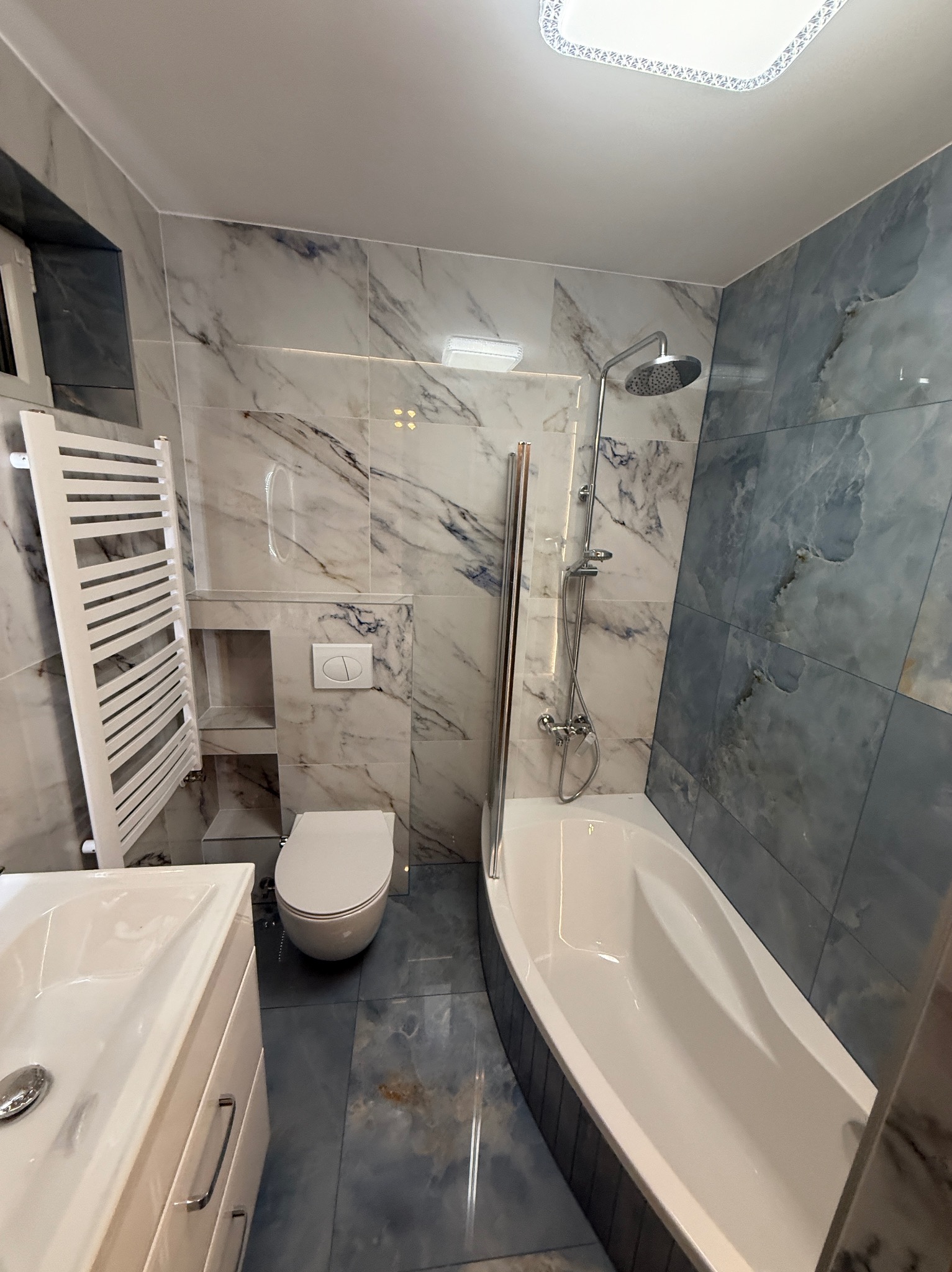 Wykończona łazienka w odcieniach bieli i błękitu z marmurkowymi płytkami, grzejnikiem drabinkowym i podtynkową spłuczką. Widok na wannę z deszczownicą.