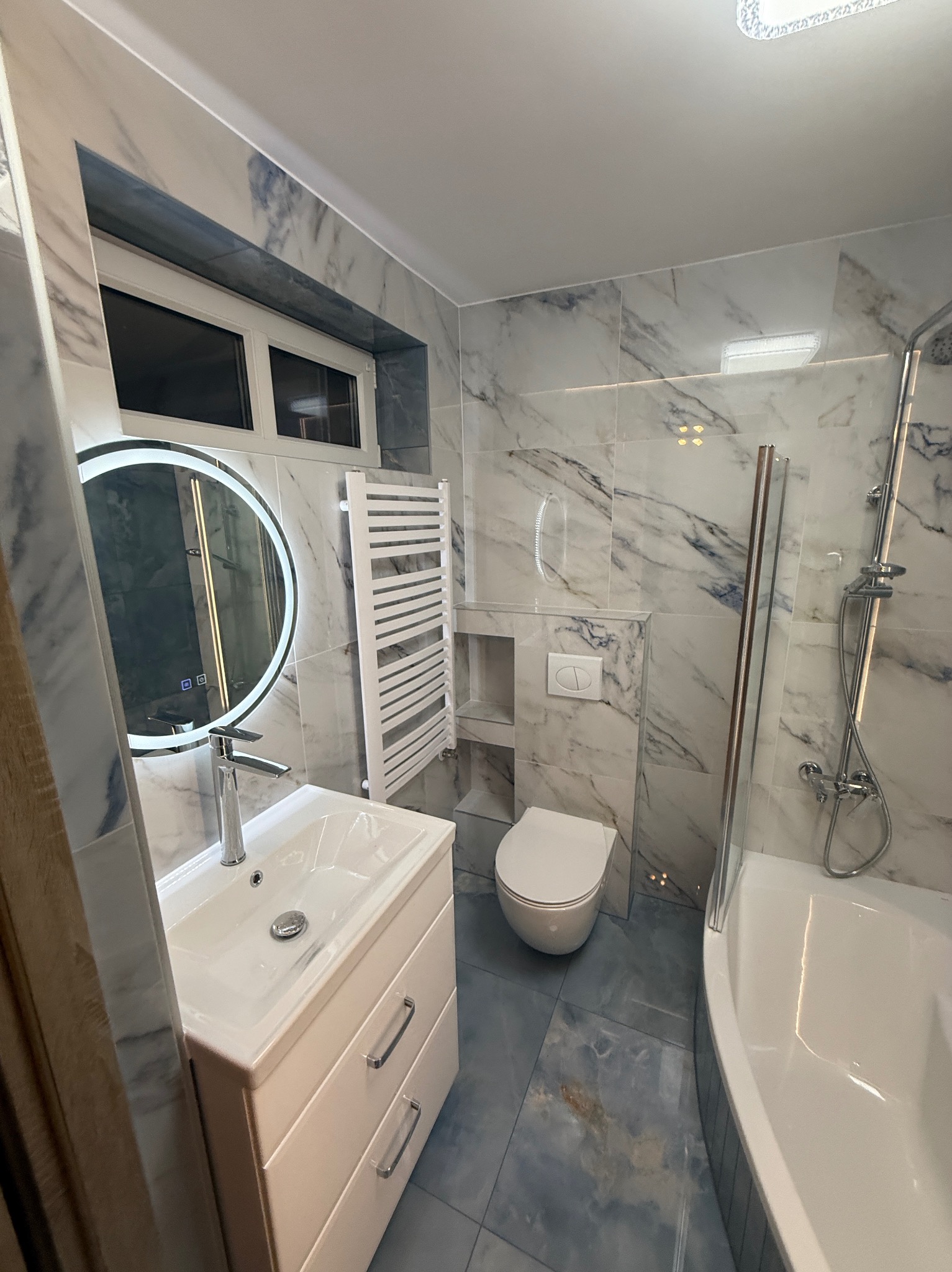 Nowoczesna łazienka z marmurowymi płytkami, okrągłym lustrem z podświetleniem LED, białą armaturą i grzejnikiem drabinkowym. Błękitna podłoga.