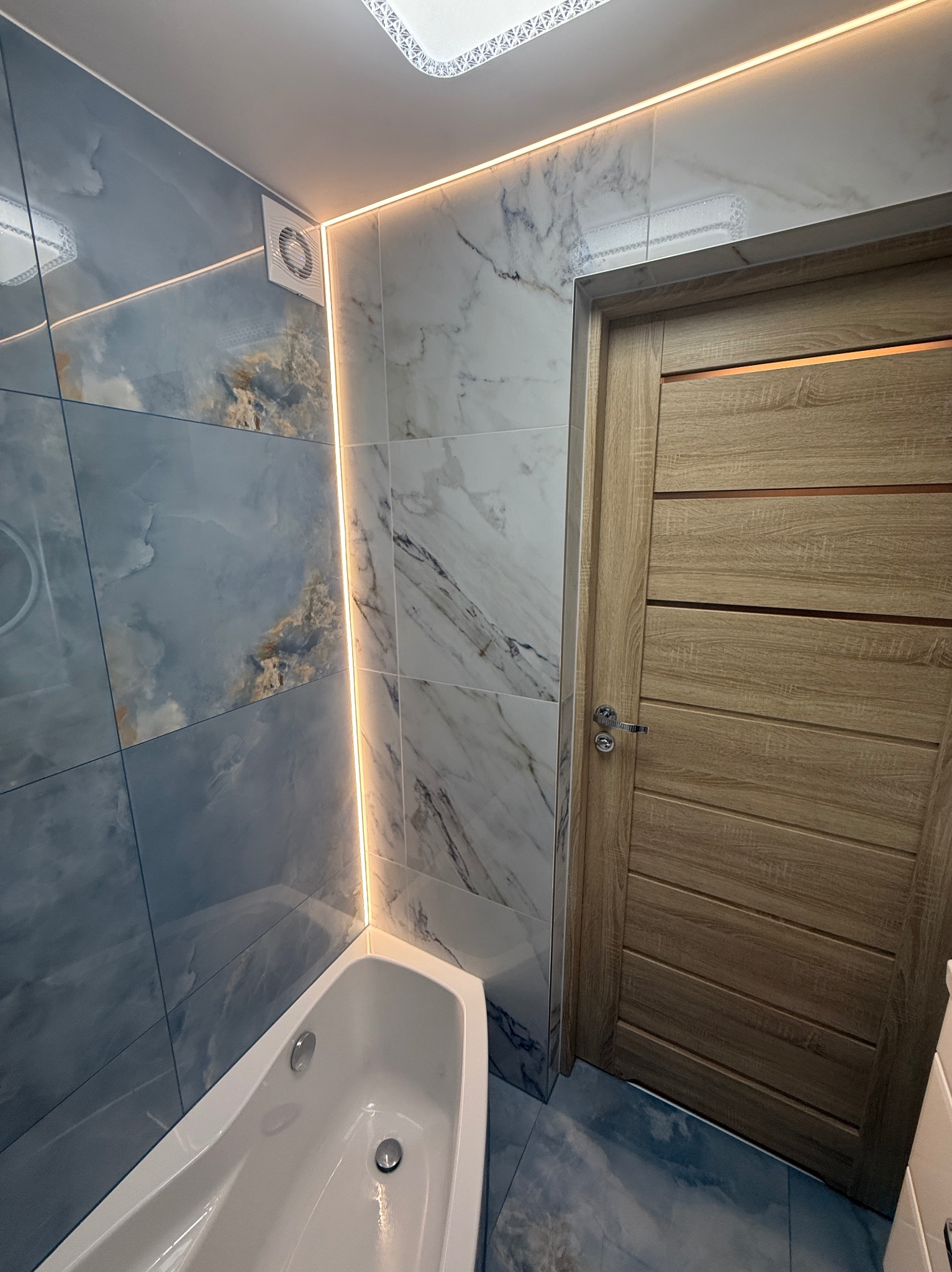 Wykończona łazienka z marmurowymi płytkami, podświetleniem LED i drewnianymi drzwiami. Widoczna wanna i wentylator.