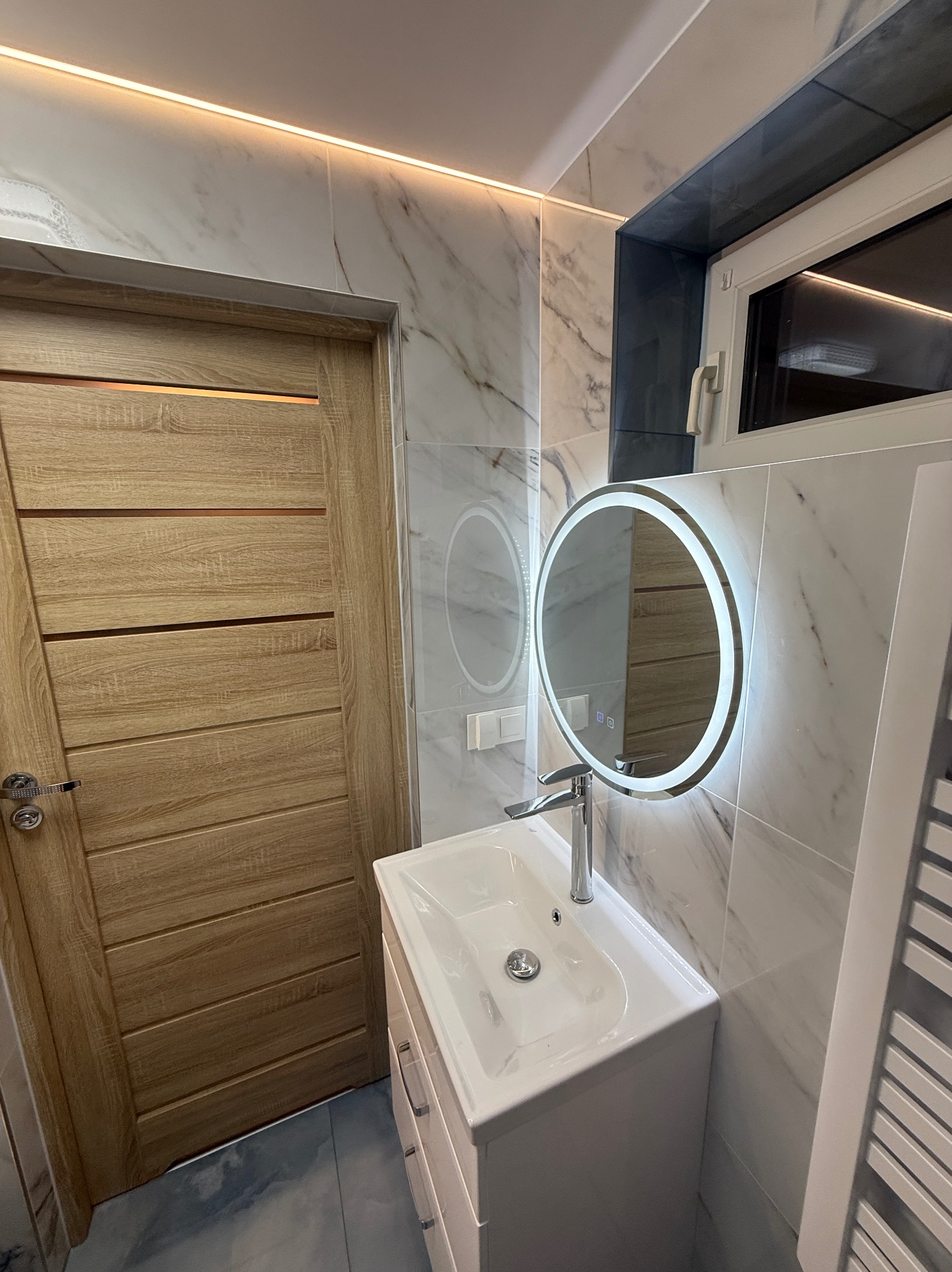 Nowoczesna łazienka: biała umywalka z szafką, okrągłe lustro LED, marmurowe płytki ścienne, drewniane drzwi i subtelne oświetlenie sufitowe.