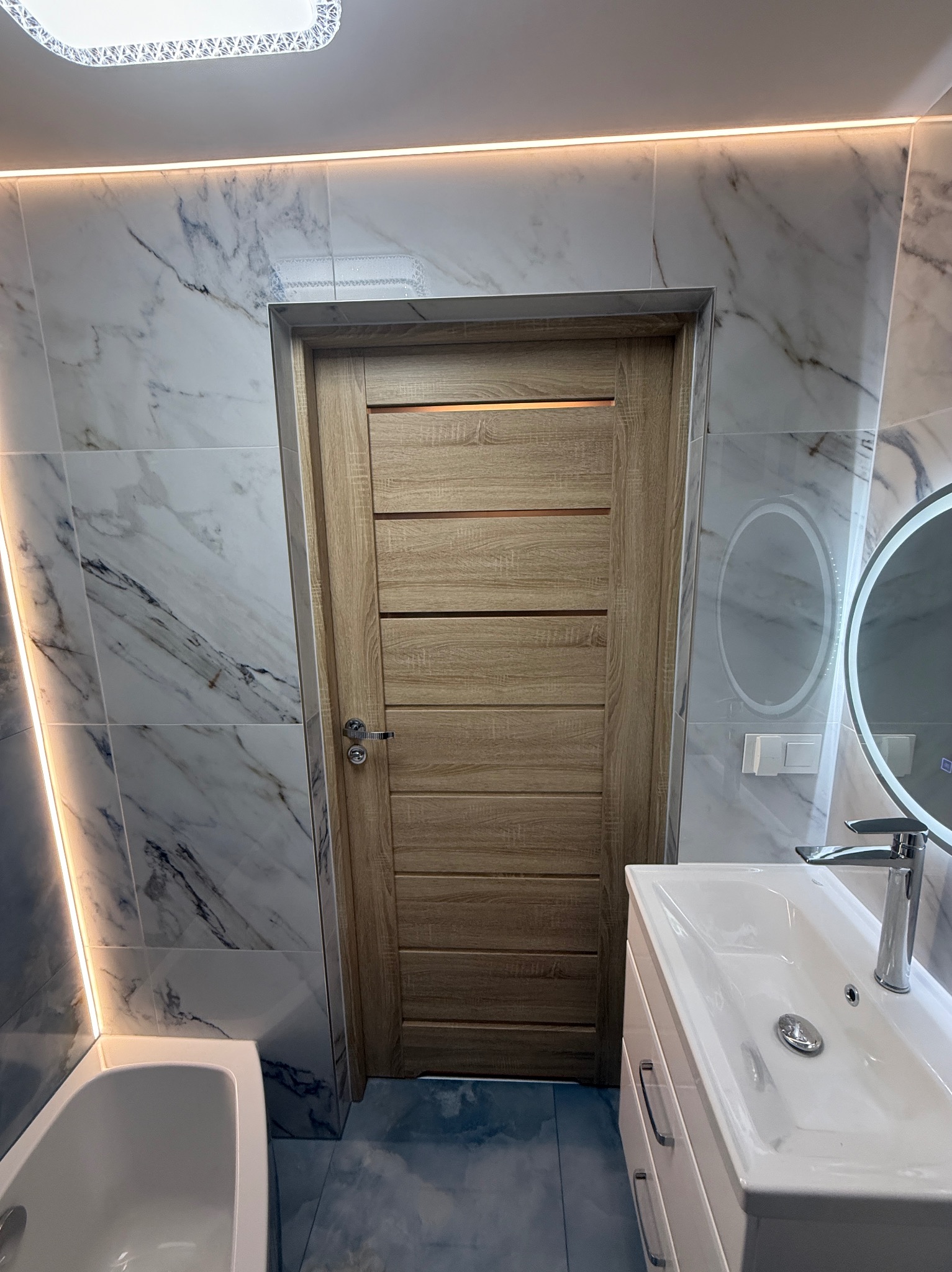 Nowoczesna łazienka z marmurowymi płytkami, drewnianymi drzwiami i okrągłym lustrem z podświetleniem LED. Umywalka z szafką i chromowaną baterią.