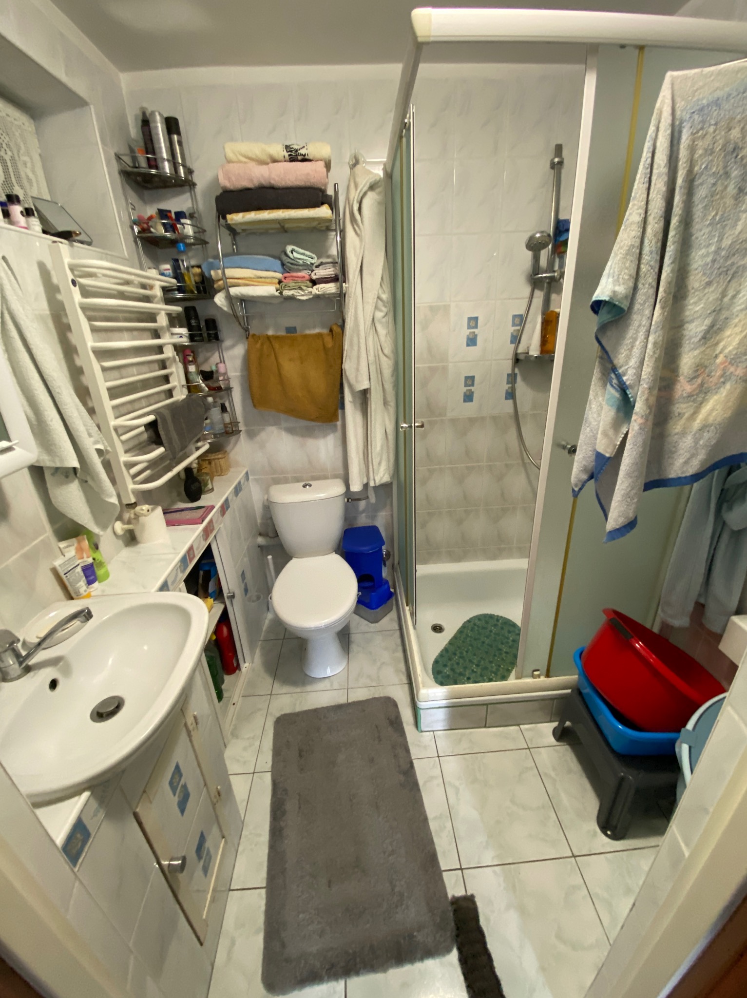 Umywalka, toaleta i kabina prysznicowa w małej, wykafelkowanej łazience. Widoczne półki z kosmetykami i ręcznikami, dywanik podłogowy. Ujęcie z perspektywy.