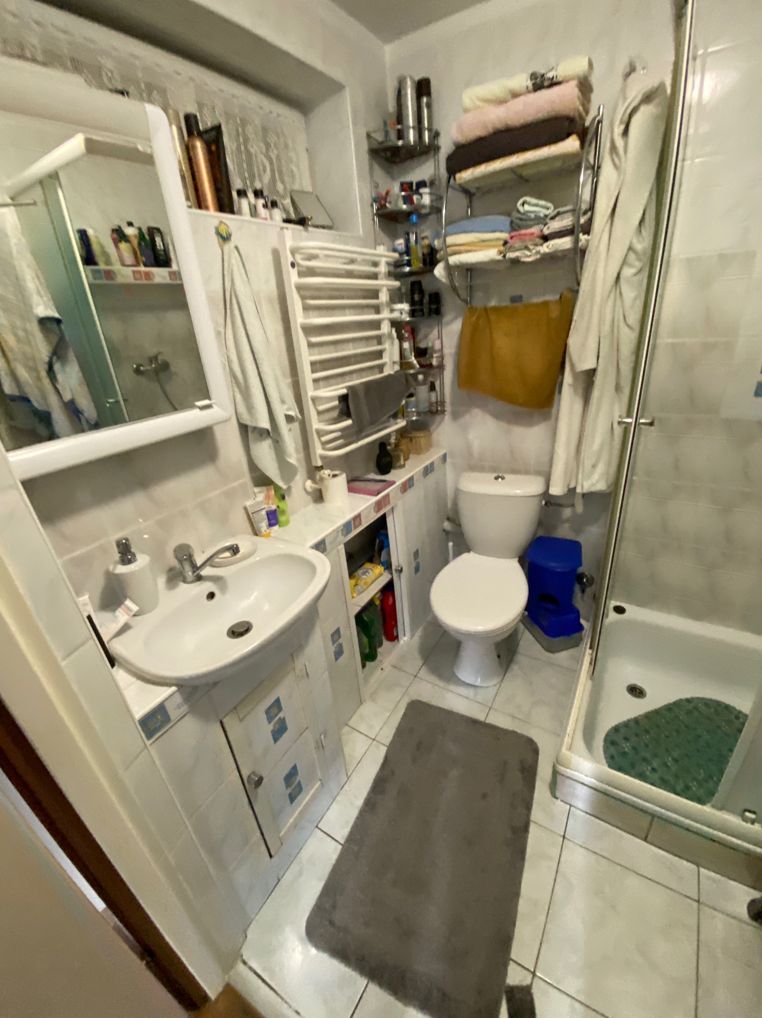 Wykończona łazienka z kabiną prysznicową, umywalką z szafką, toaletą i grzejnikiem drabinkowym. Widoczne płytki ceramiczne i akcesoria łazienkowe.