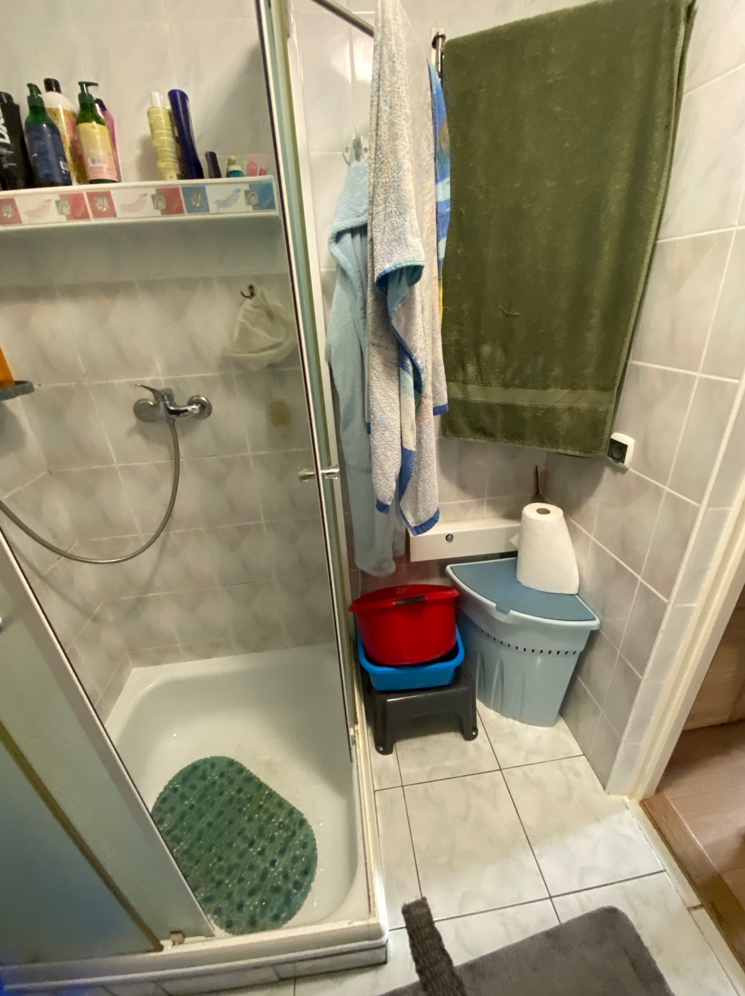 Wnętrze łazienki z kabiną prysznicową, na półce kosmetyki, obok ręczniki i akcesoria łazienkowe. Widoczne płytki na ścianach i podłodze.