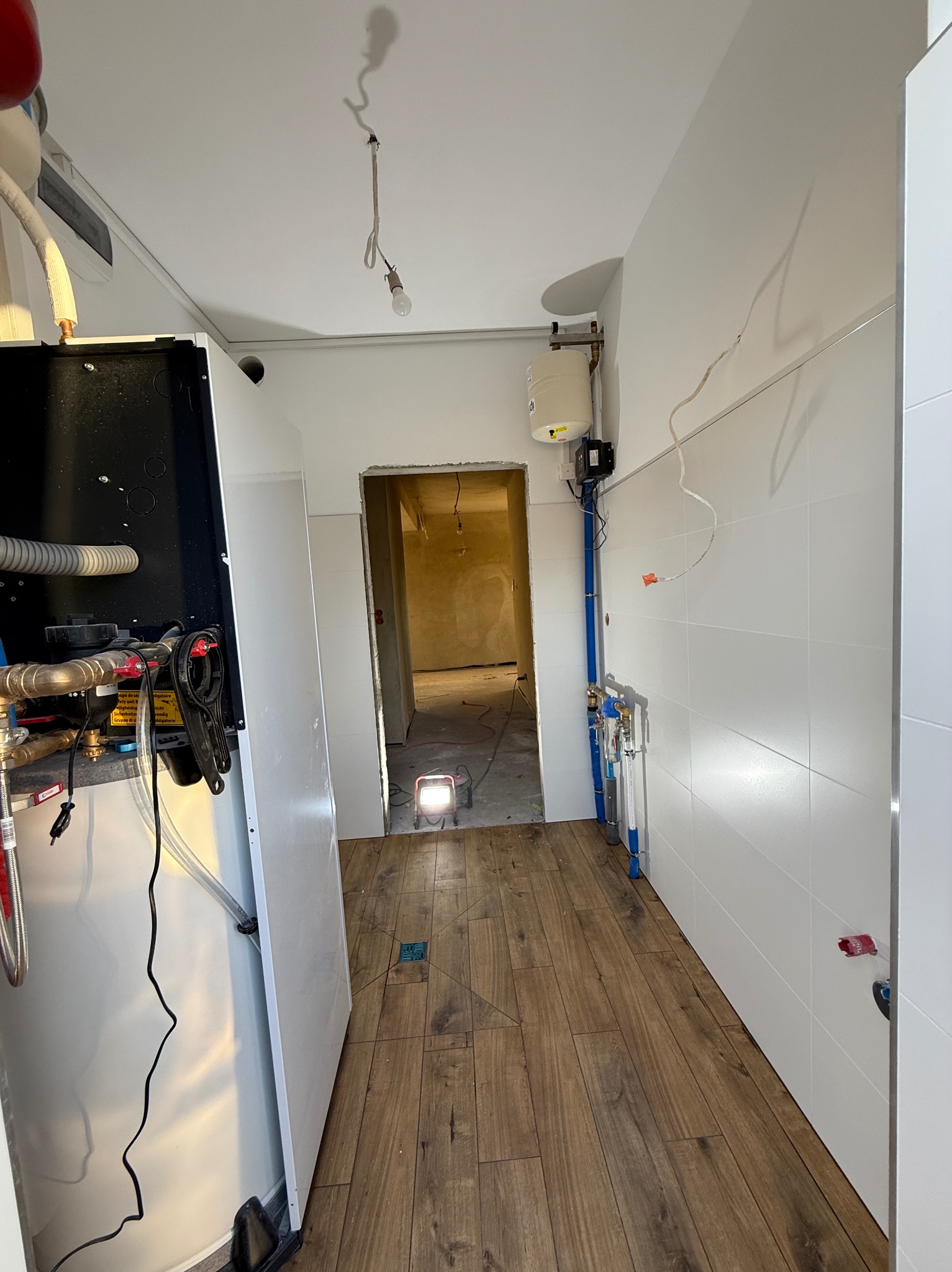 Wnętrze łazienki w trakcie prac: białe płytki na ścianach, drewnopodobna podłoga, instalacja wodna z niebieskich rur. W głębi widoczne surowe ściany innego pomieszczenia.