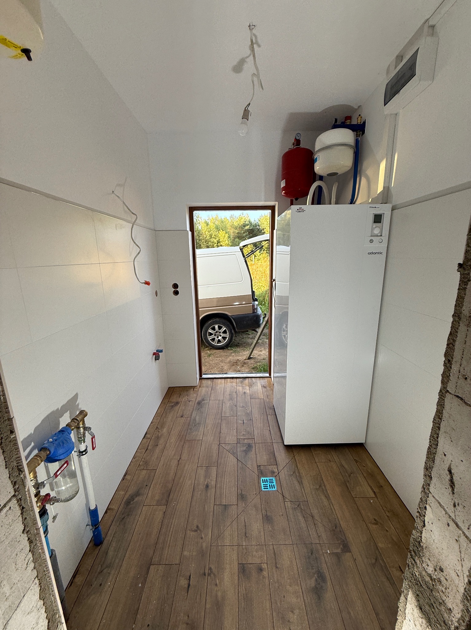 Wykończona łazienka z drewnopodobną podłogą i białymi płytkami. Widoczny bojler, instalacja wodna i częściowo samochód dostawczy w otwartych drzwiach.