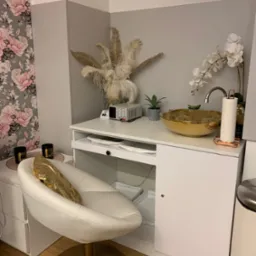 Nowoczesne stanowisko do manicure z białym fotelem, złotą umywalką i dekoracjami w salonie kosmetycznym, widoczne akcesoria i sprzęt.