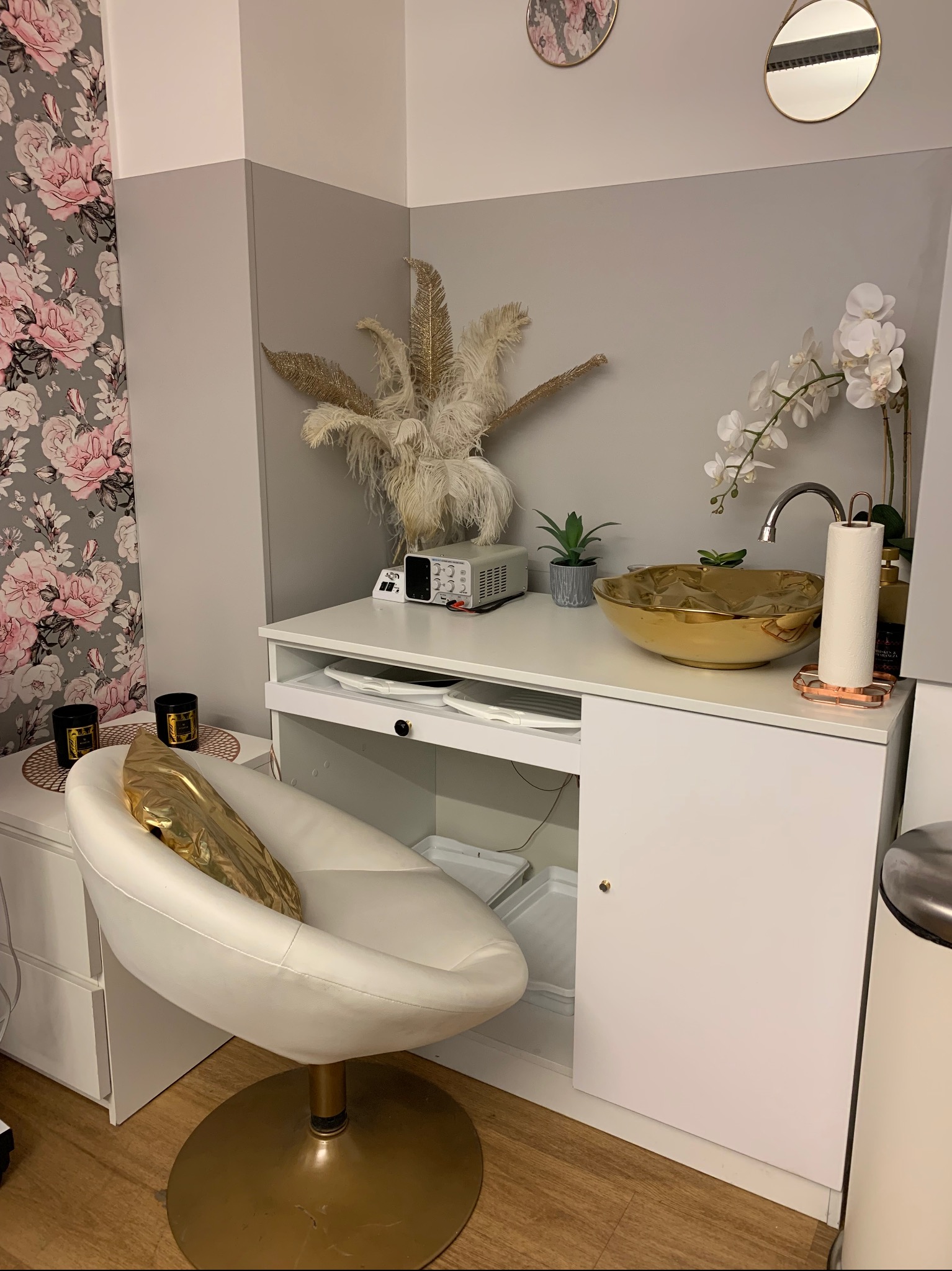 Nowoczesne stanowisko do manicure z białym fotelem, złotą umywalką i dekoracjami w salonie kosmetycznym, widoczne akcesoria i sprzęt.