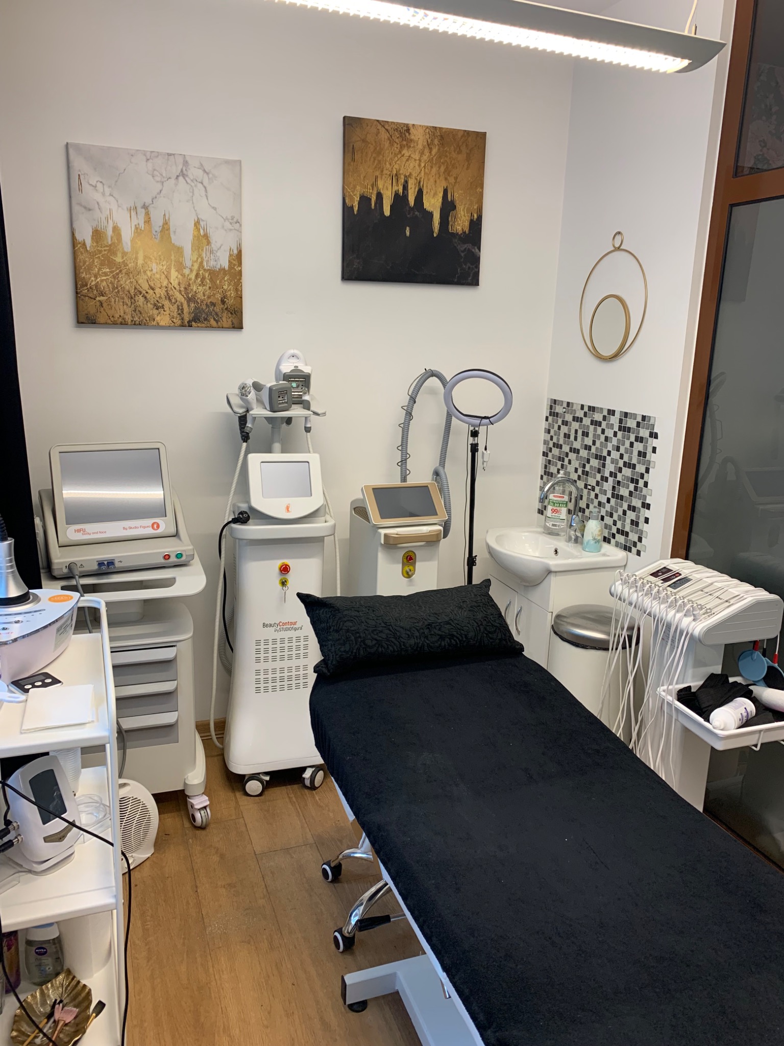 Wnętrze gabinetu z leżanką kosmetyczną, urządzeniami do zabiegów, umywalką i dekoracjami ściennymi w postaci obrazów i lustra.