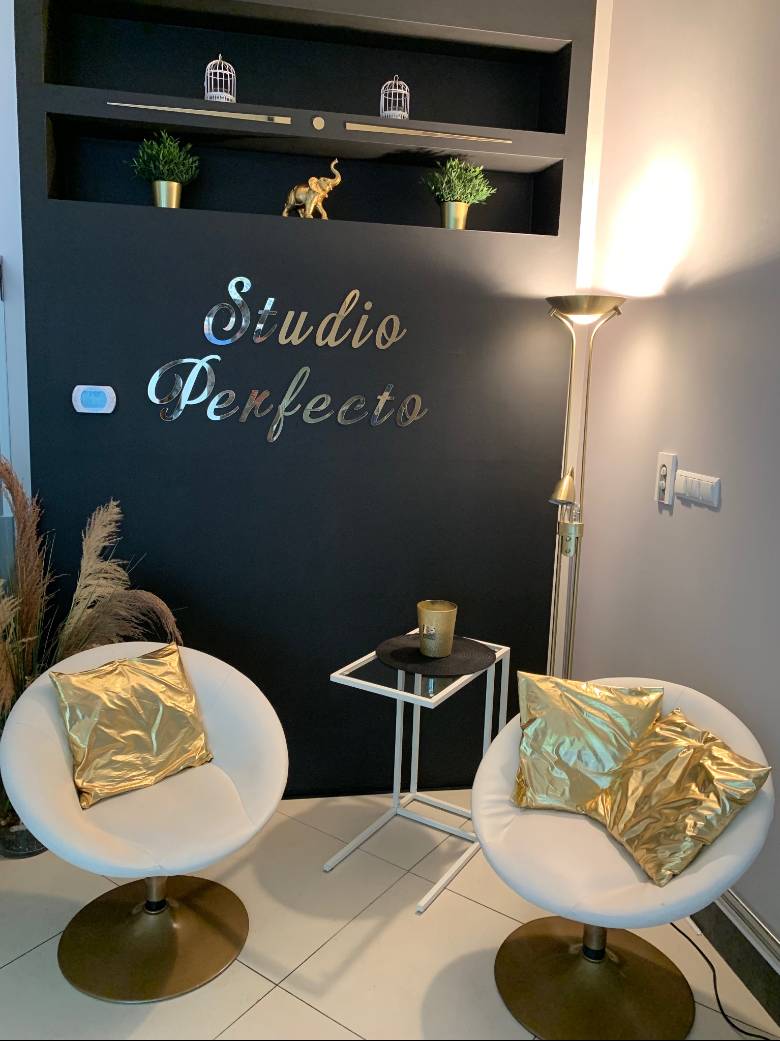 Elegancki kącik poczekalni w salonie z dwoma białymi fotelami z złotymi poduszkami, stolikiem i lampą podłogową, na czarnej ścianie srebrny napis 'Studio Perfecto' i dekoracyjne półki z ozdobami.