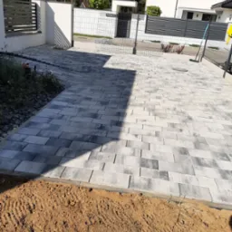 Nowoczesny podjazd z szarej kostki brukowej ułożonej w regularny wzór, obrzeża z ziemi przygotowane pod dalsze prace, widoczny fragment ogrodzenia i domu w tle.