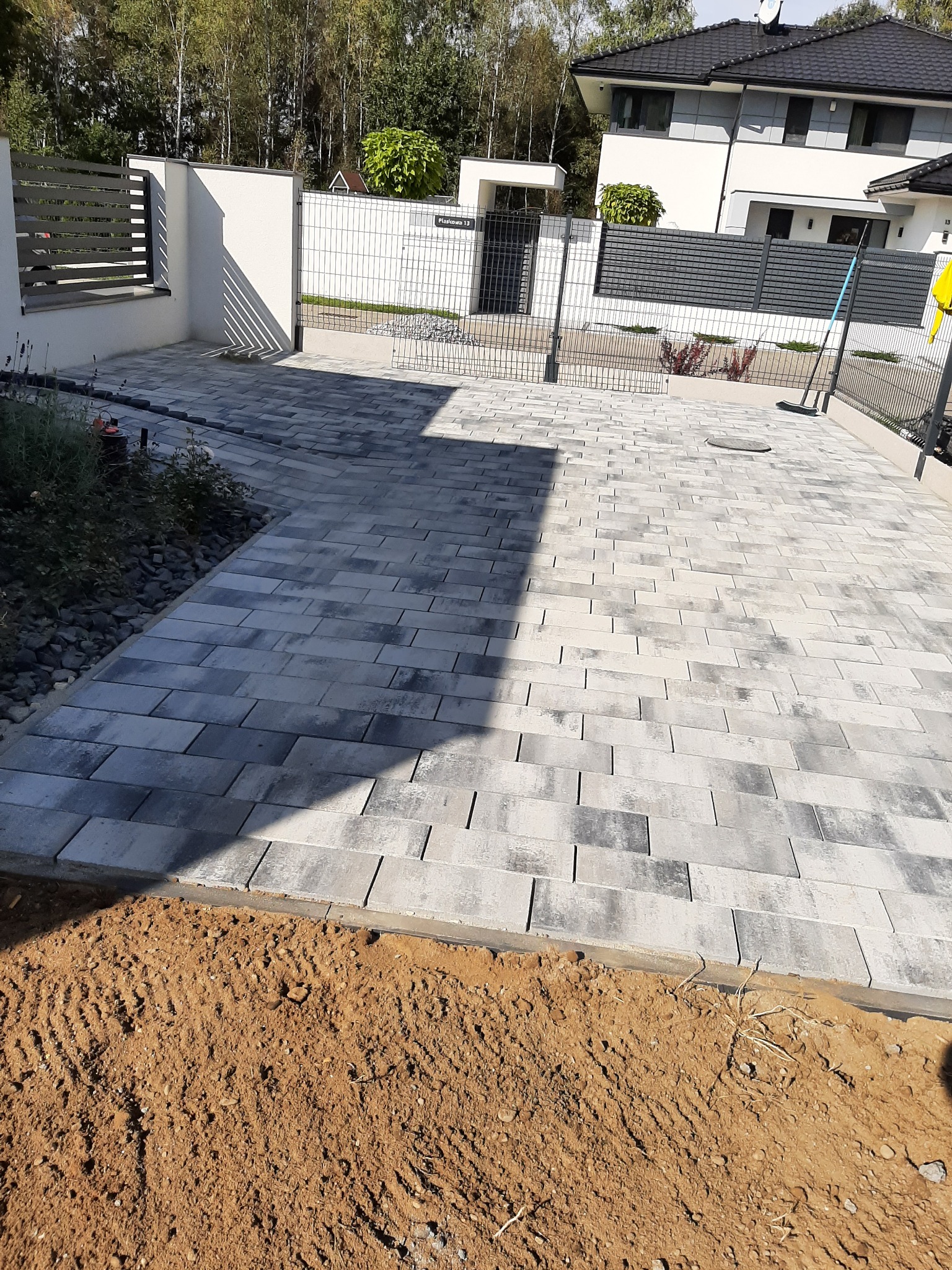 Nowoczesny podjazd z szarej kostki brukowej ułożonej w regularny wzór, obrzeża z ziemi przygotowane pod dalsze prace, widoczny fragment ogrodzenia i domu w tle.