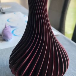 Kamil Kol - Burgundowa, spiralna waza z drewna lub tworzywa, widoczna na stole z niebieskim elementem dekoracyjnym w tle.