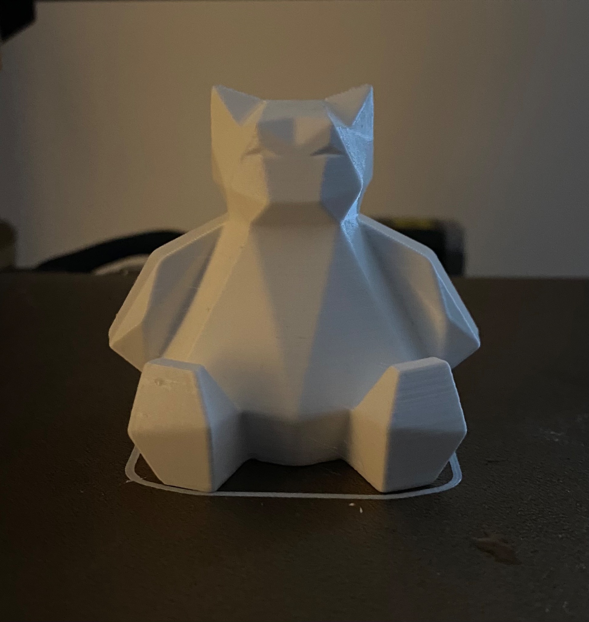 Biała, geometryczna figurka kota wykonana w technologii druku 3D, widoczne warstwy druku, na ciemnym tle.