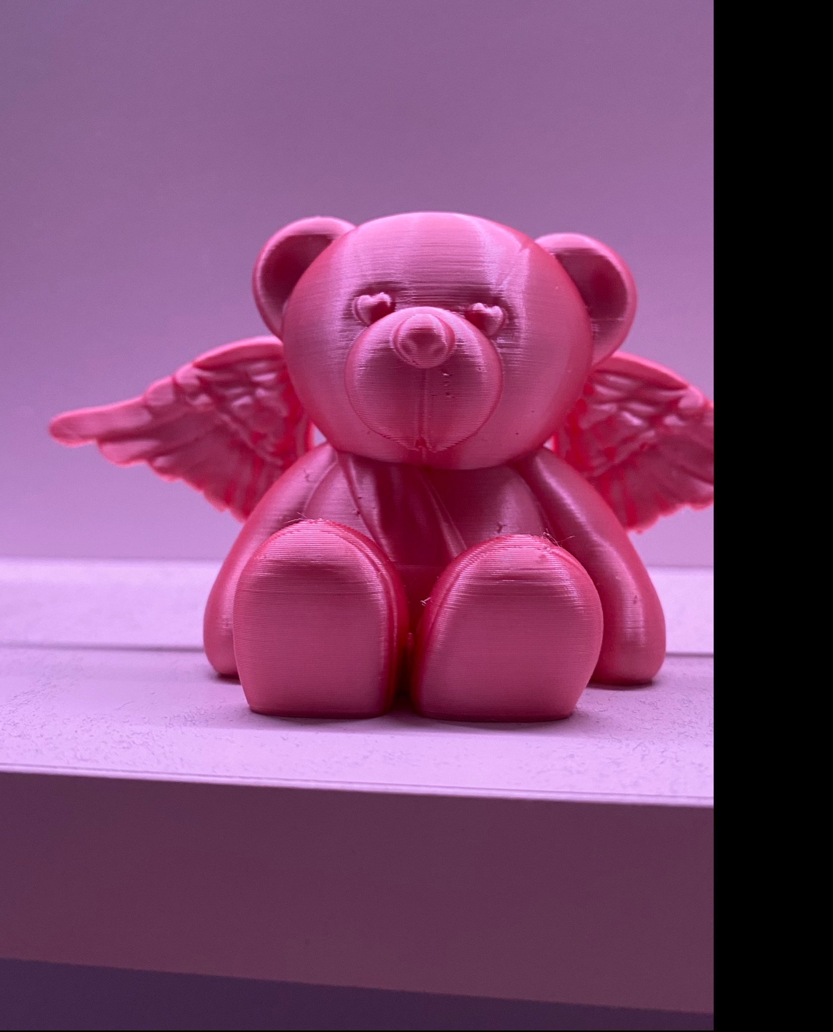 Różowy, drukowany w 3D miś z małymi skrzydłami anioła i oczami w kształcie serc, siedzący na jasnej powierzchni pod delikatnym, różowym oświetleniem.