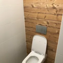 Nowoczesna toaleta podwieszana z deską wolnoopadającą, obudowana płytkami imitującymi drewno i białymi płytkami ceramicznymi, widoczny przycisk spłuczki w kolorze srebrnym.