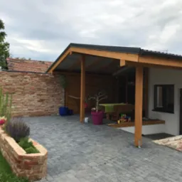 Nowoczesna altana z drewnianą konstrukcją i zadaszeniem, widok na brukowany podjazd, ceglany murek z kwiatami i zielony trawnik w tle.