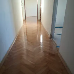 Jasny, parkietowy korytarz ułożony w jodełkę, świeżo po lakierowaniu, z widocznymi listwami przypodłogowymi i przygotowanymi do zabezpieczenia elementami w przejściu.