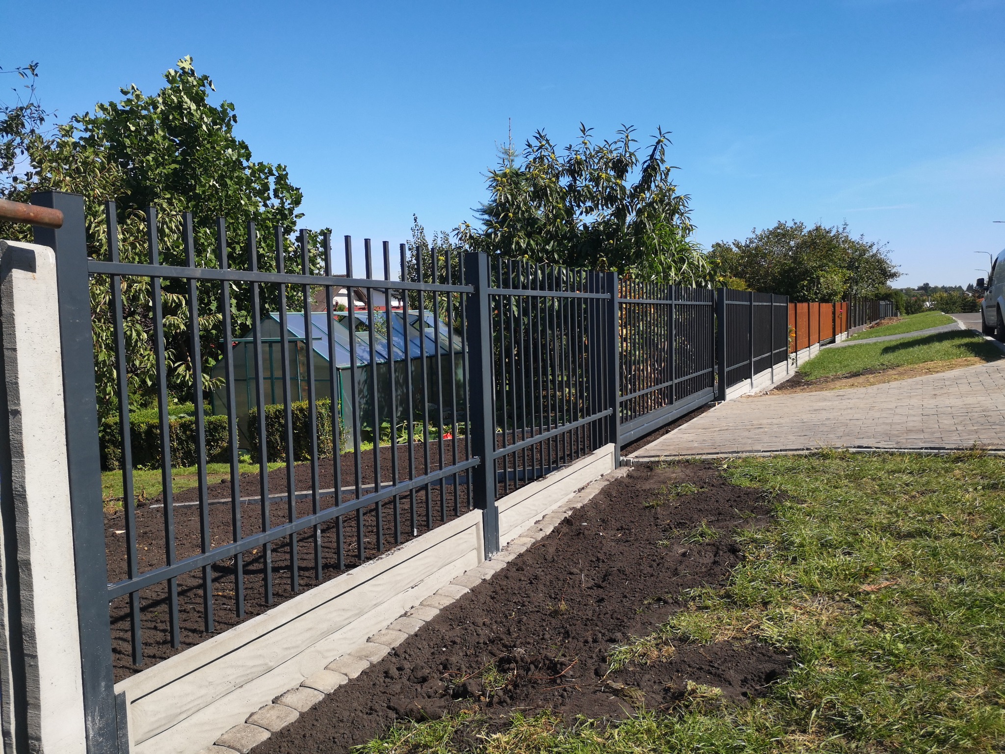 Nowoczesne, grafitowe ogrodzenie panelowe z betonową podmurówką i obrzeżem z kostki, na tle zieleni i błękitnego nieba.