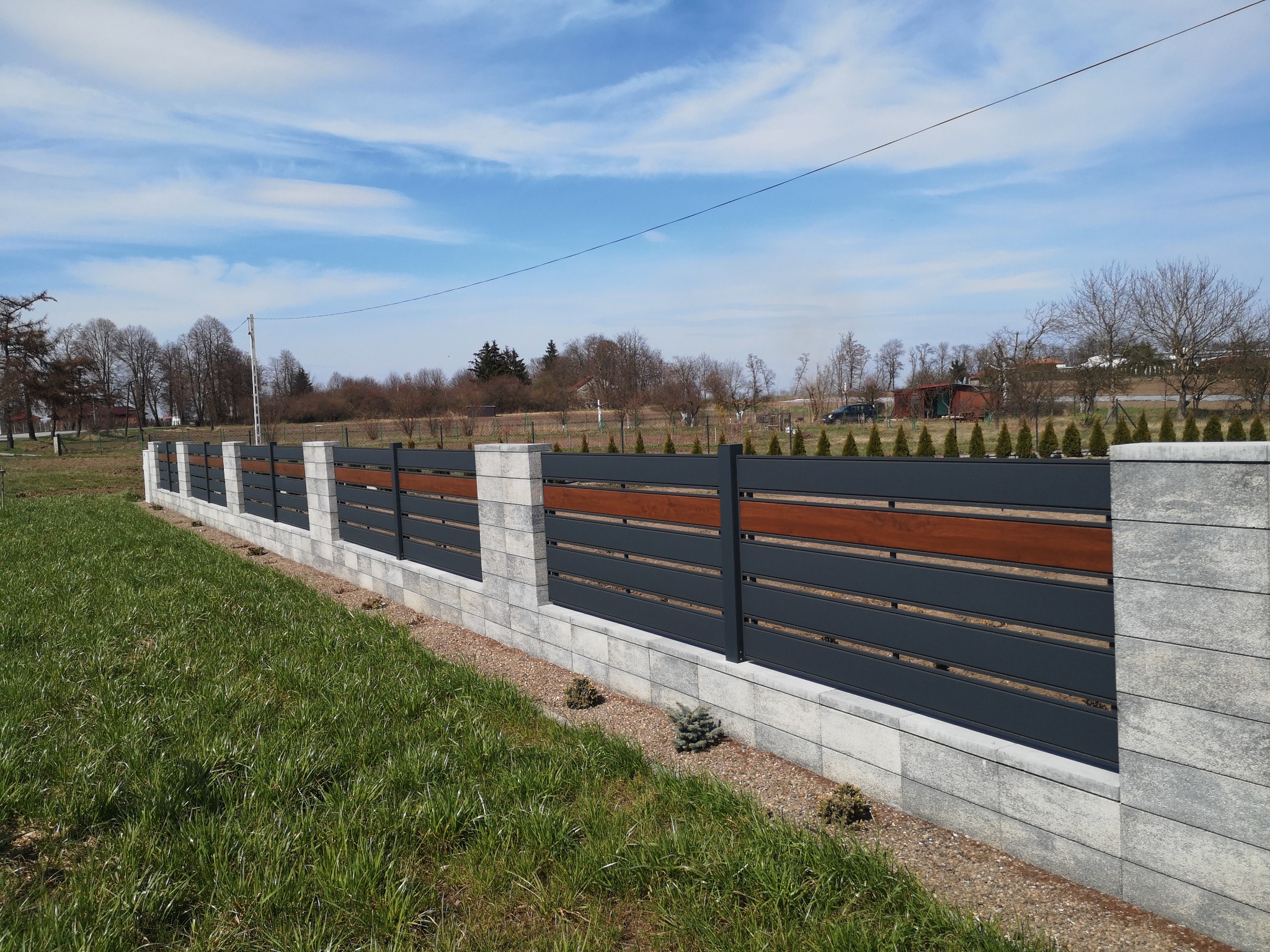 Nowoczesne ogrodzenie panelowe z elementami drewnianymi i kamiennymi słupkami, oddzielające trawnik od posesji.