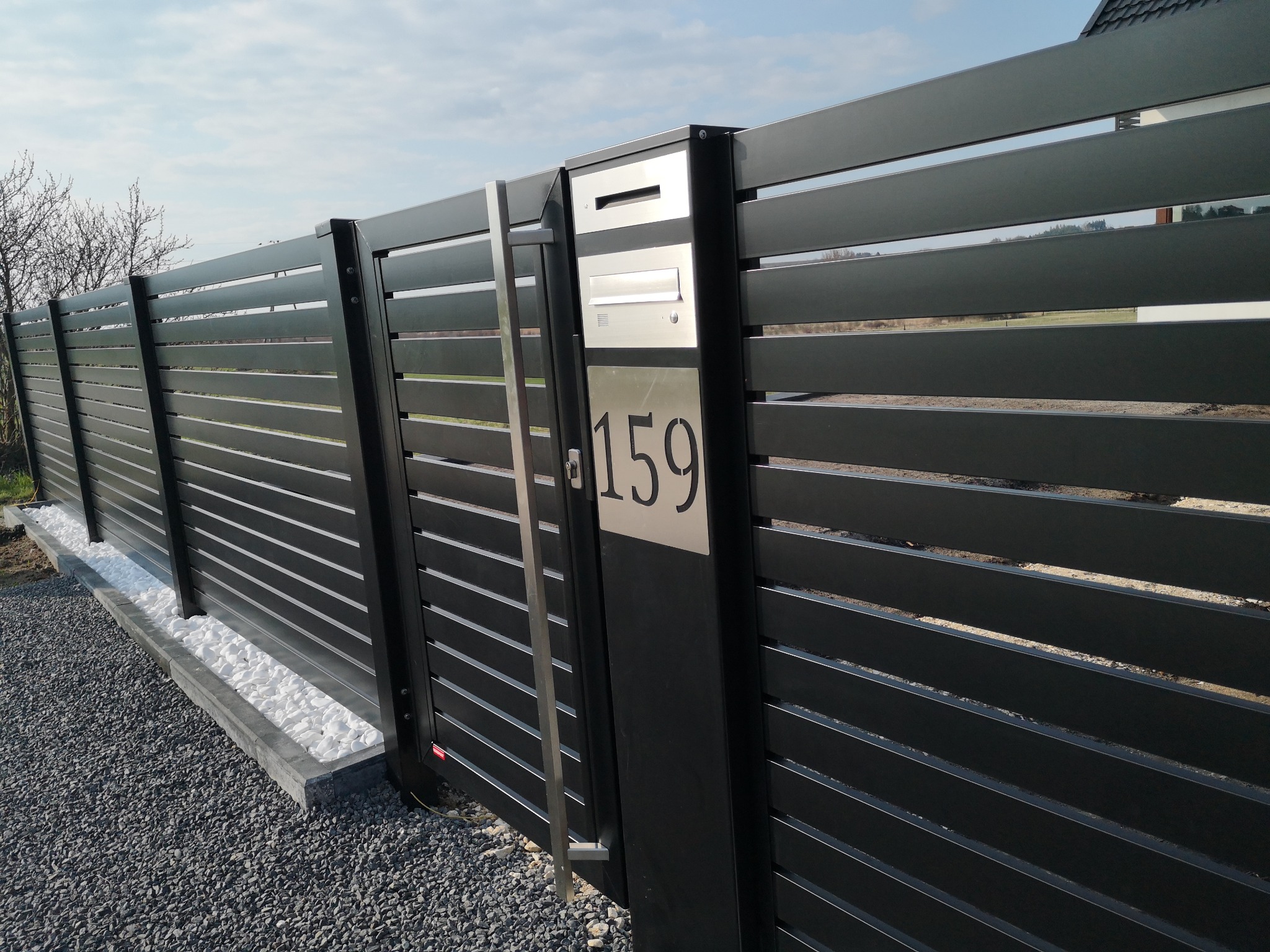 Nowoczesne, poziome, czarne ogrodzenie z metalu z numerem domu 159 i skrzynką na listy, otoczone białymi kamieniami i szarym żwirem.