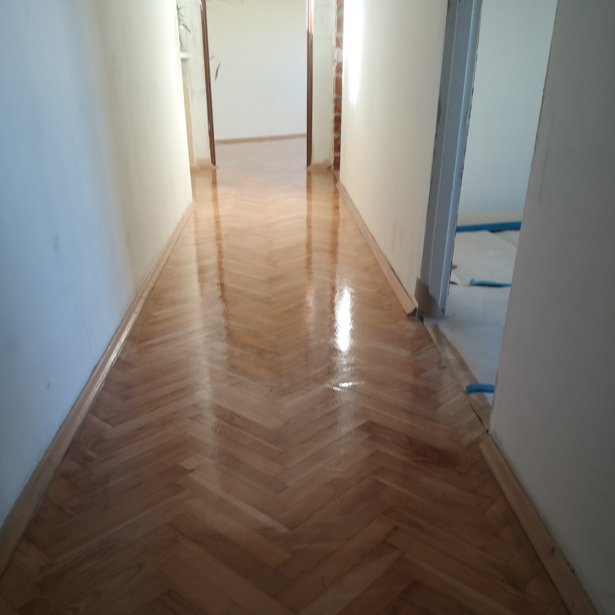 Jasny, parkietowy korytarz ułożony w jodełkę, świeżo po lakierowaniu, z widocznymi listwami przypodłogowymi i przygotowanymi do zabezpieczenia elementami w przejściu.