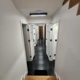 Widok z góry na korytarz z czarną marmurową podłogą i rzędem białych drzwi z czarnymi klamkami, widoczne drewniane schody w dolnej części kadru, oświetlenie sufitowe LED.