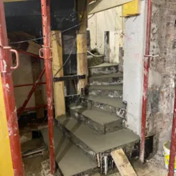 Wylewanie betonowych schodów spiralnych w trakcie remontu, widoczne podpory i szalunki, surowe ściany w tle.