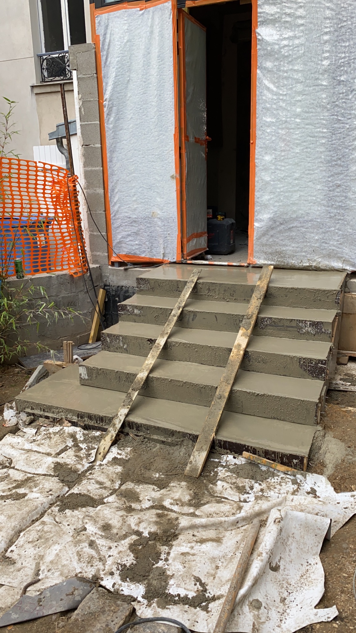 Nowe, betonowe schody wejściowe w trakcie budowy, z deskami podtrzymującymi, przed zabezpieczonymi folią drzwiami.