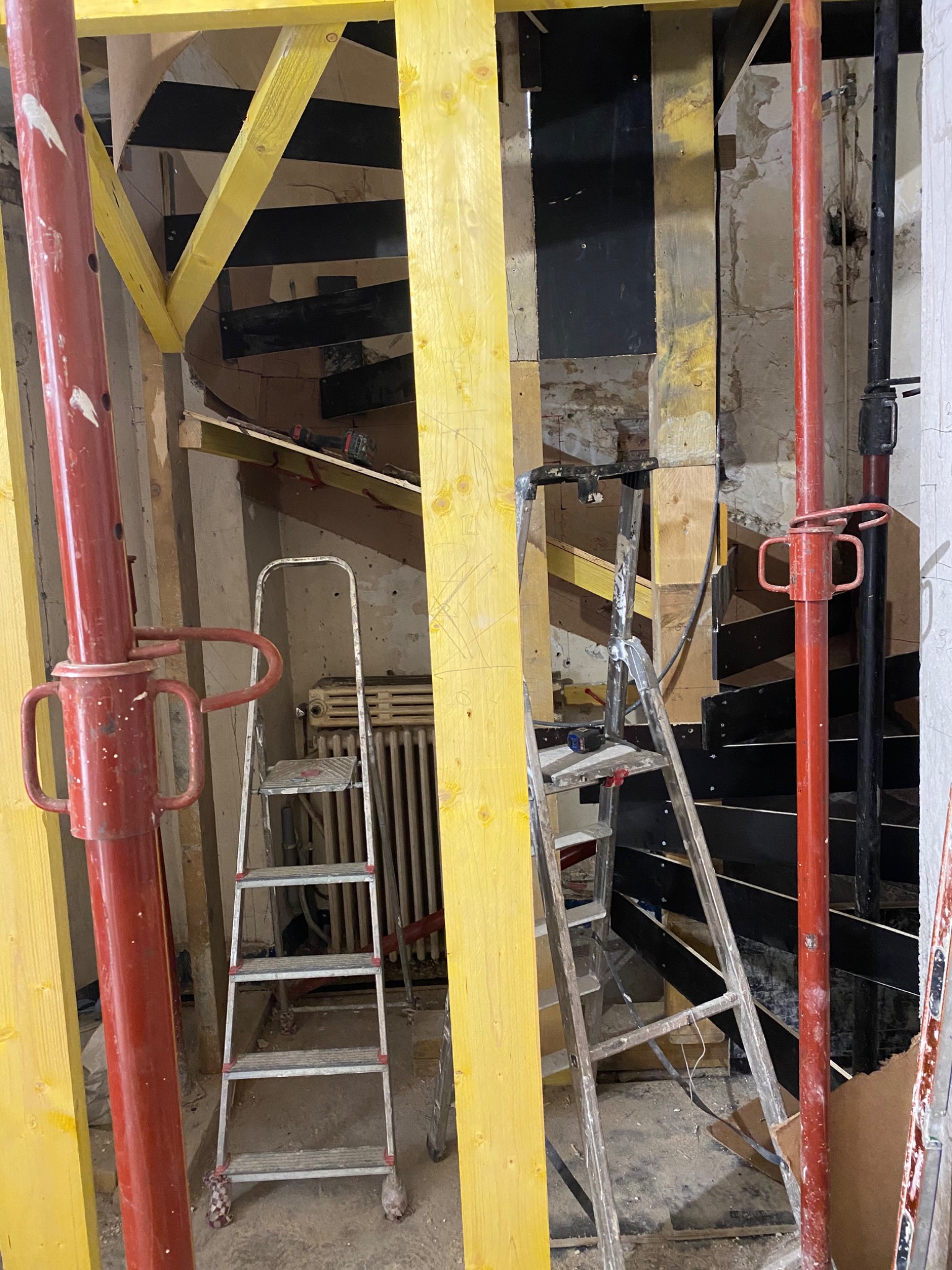 Wnętrze w trakcie remontu ze schodami: widoczne podpory budowlane, drabiny aluminiowe, elementy drewniane konstrukcji schodów oraz fragment starego grzejnika.