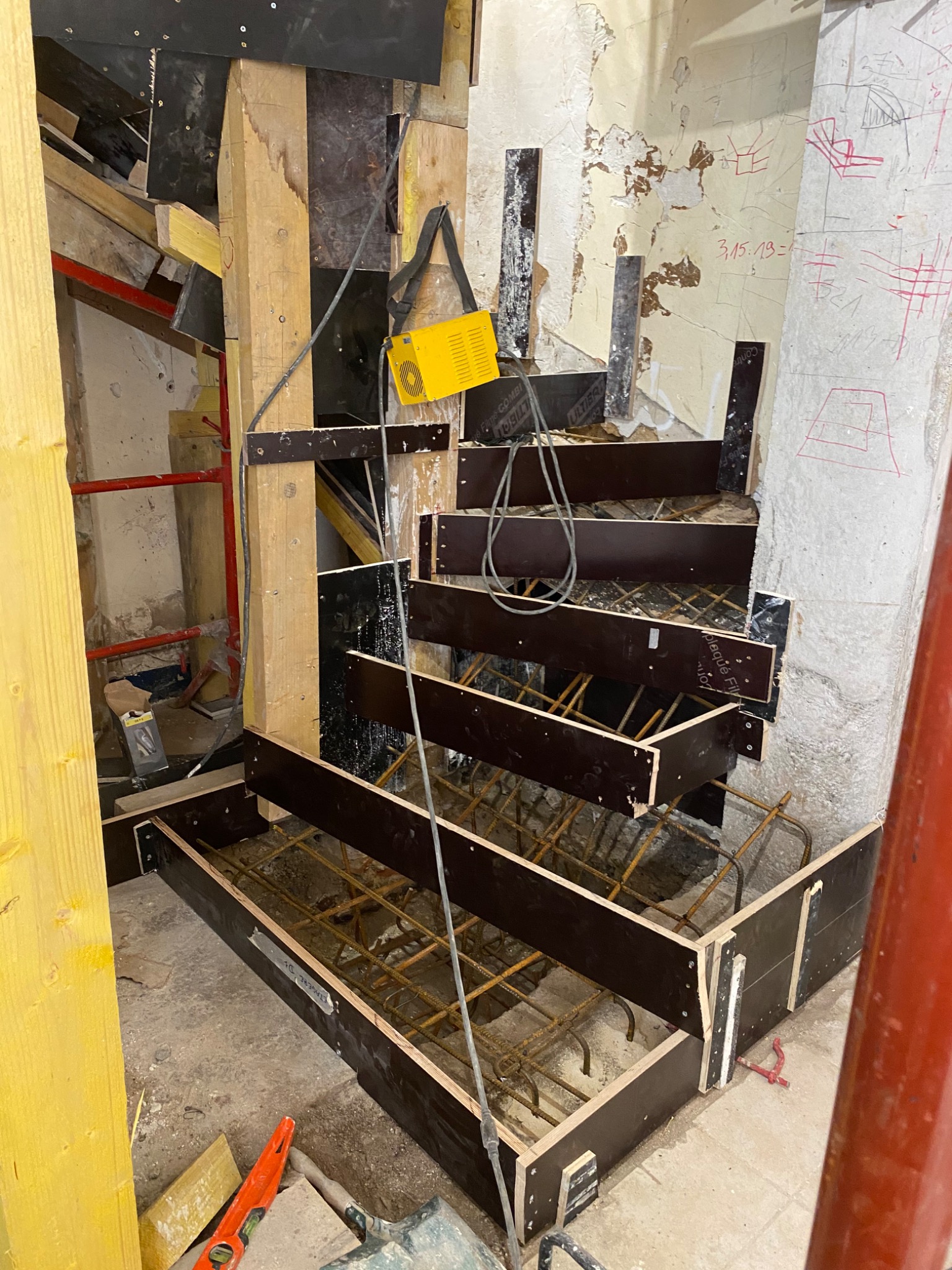 Szare schody w trakcie budowy, z widocznym zbrojeniem i szalunkiem z drewnianych desek, na tle starej ściany z resztkami farby i notatkami.