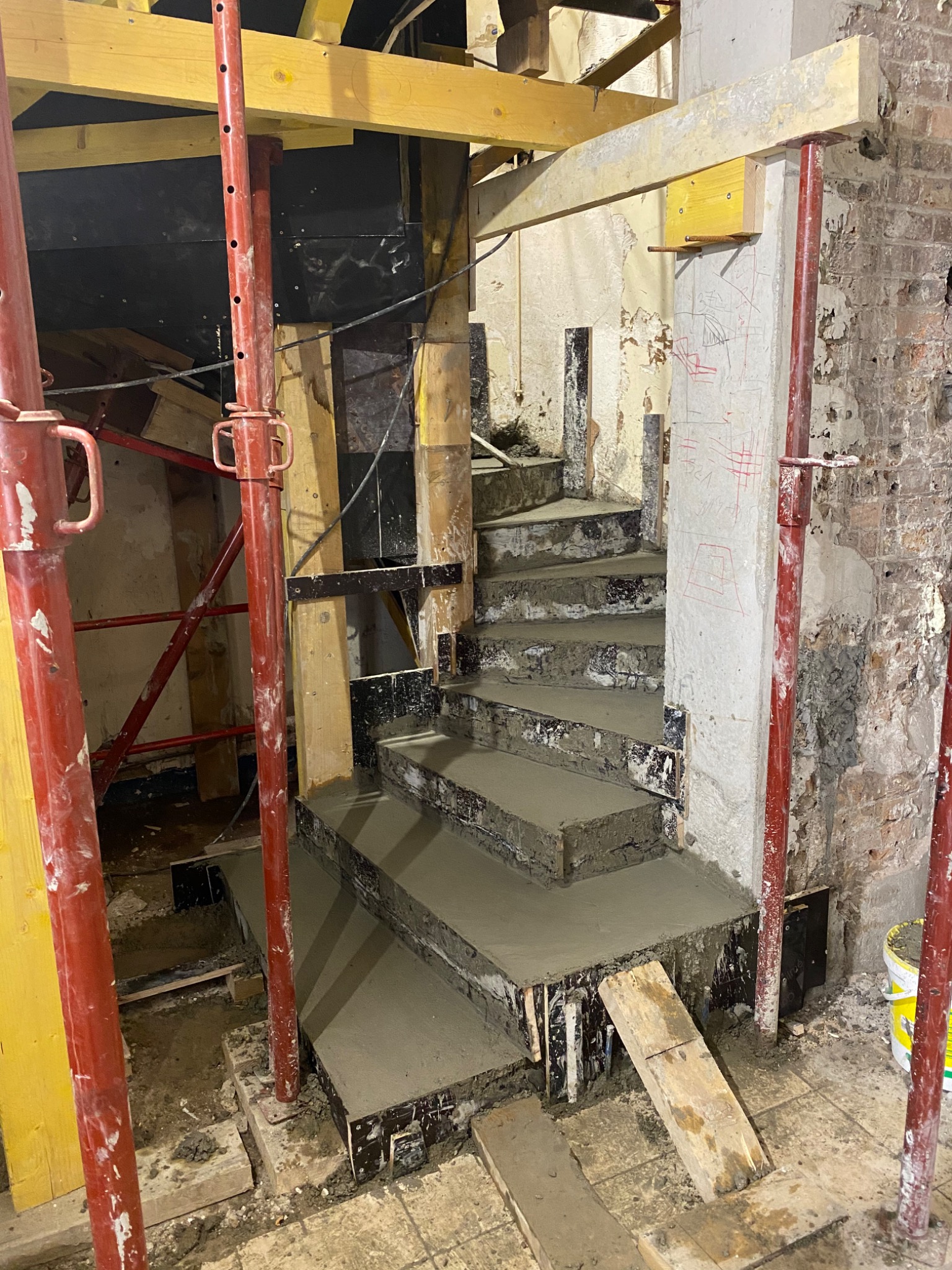 Wylewanie betonowych schodów spiralnych w trakcie remontu, widoczne podpory i szalunki, surowe ściany w tle.