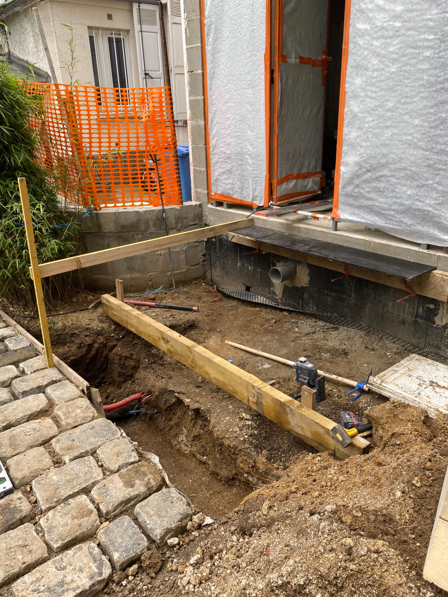 Przygotowanie wykopu pod fundament budynku z zabezpieczeniem wykopu drewnianymi belkami, widoczne narzędzia budowlane i pomarańczowa siatka zabezpieczająca.