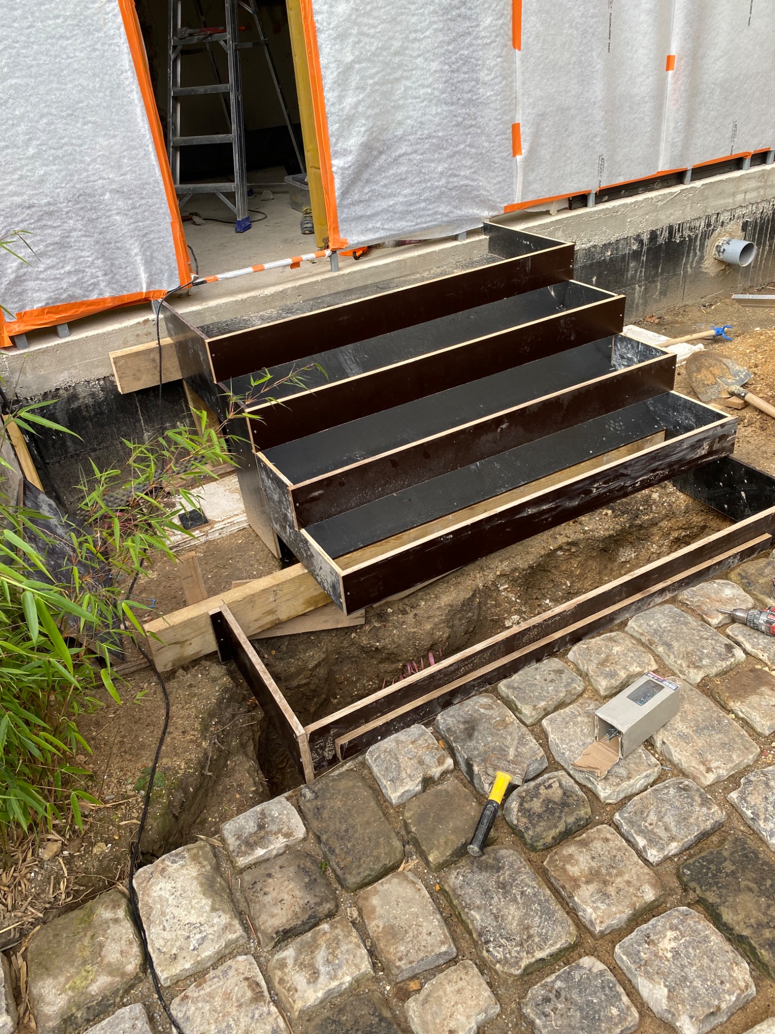 Budowa schodów zewnętrznych z drewnianym szalunkiem i kamienną kostką brukową, widoczne narzędzia i częściowo wykopana ziemia.