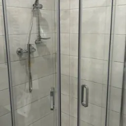 Kabina prysznicowa z chromowaną armaturą i szarymi, kwadratowymi płytkami na ścianach. Widoczne ślady użytkowania na dolnej części kabiny.