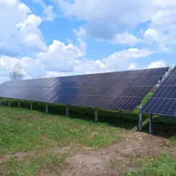 Rząd paneli słonecznych na metalowych wspornikach, zainstalowanych na zielonej trawie pod błękitnym niebem z obłokami, widoczne drzewa w tle.