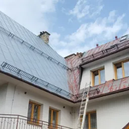 Metalowy dach po renowacji z zamontowanymi zabezpieczeniami przeciwśniegowymi, drabina oparta o ścianę budynku, widoczny fragment elewacji z kamienia i drewniane okna.