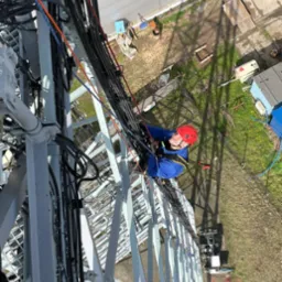 Pracownik w czerwonym kasku zabezpieczony linami, na wysokości, montuje kable na metalowej konstrukcji wieży telekomunikacyjnej, widok z góry na teren z trawą i budynkami.