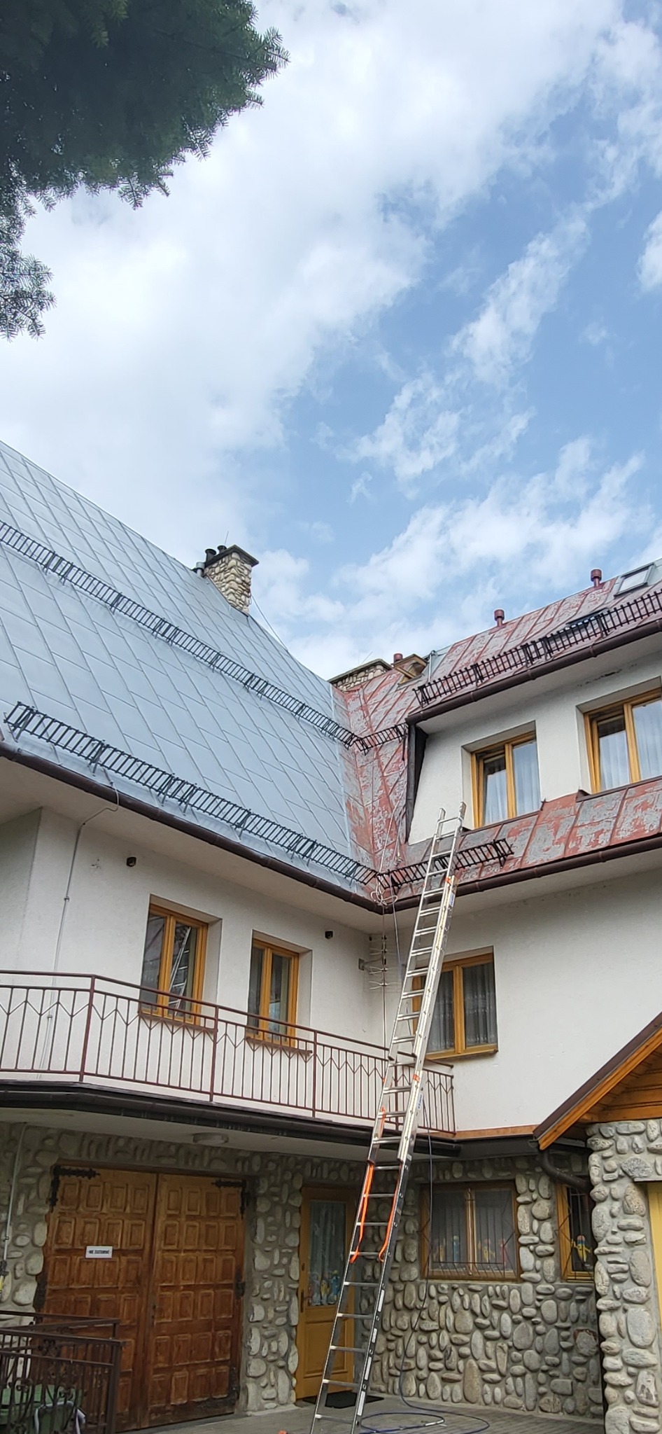 Metalowy dach po renowacji z zamontowanymi zabezpieczeniami przeciwśniegowymi, drabina oparta o ścianę budynku, widoczny fragment elewacji z kamienia i drewniane okna.