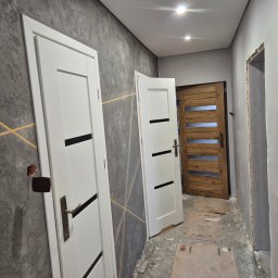 Artur Walusiewicz - Korytarz po remoncie: szara ściana z dekoracyjną fakturą i złotymi liniami, białe drzwi z czarnymi akcentami, drewniane drzwi w głębi. Widoczne ślady prac wykończeniowych.