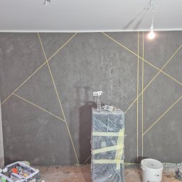Artur Walusiewicz - Dekoracyjna ściana w odcieniach szarości z geometrycznym wzorem w złotych liniach. Widoczne narzędzia malarskie i wiadro z farbą. Wykończone wnętrze.