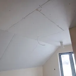 Skośny sufit poddasza wykończony płytami karton gips, widoczne łączenia płyt, okno dachowe z białym parapetem oraz wystający przewód elektryczny.