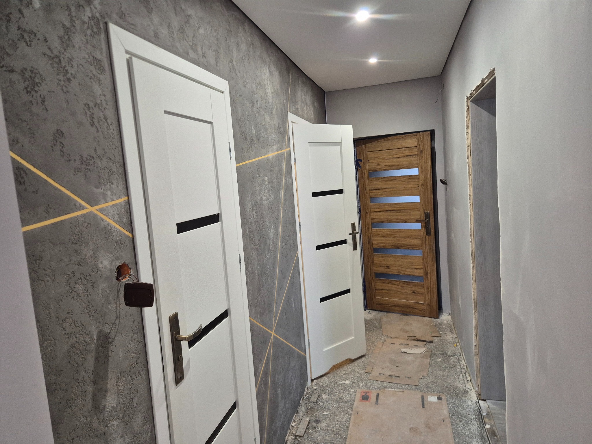 Korytarz po remoncie: szara ściana z dekoracyjną fakturą i złotymi liniami, białe drzwi z czarnymi akcentami, drewniane drzwi w głębi. Widoczne ślady prac wykończeniowych.