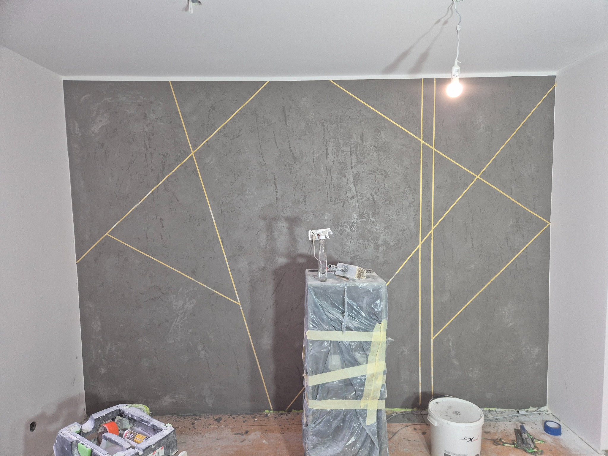 Dekoracyjna ściana w odcieniach szarości z geometrycznym wzorem w złotych liniach. Widoczne narzędzia malarskie i wiadro z farbą. Wykończone wnętrze.