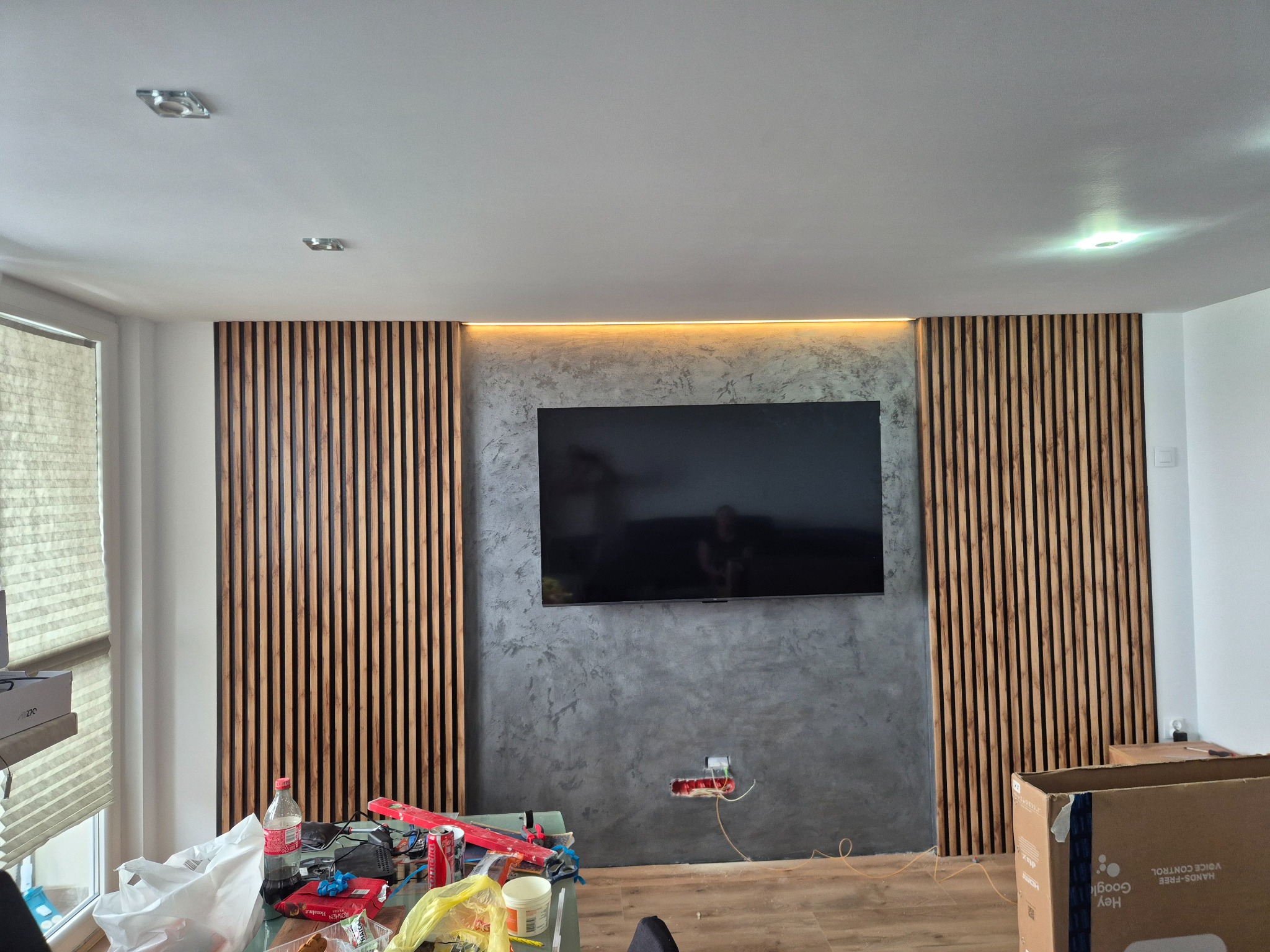 Ściana z dekoracyjnymi lamelami drewnianymi po bokach i szarą strukturą podświetloną LED nad telewizorem. Widoczne gniazdko elektryczne i podłoga z paneli.