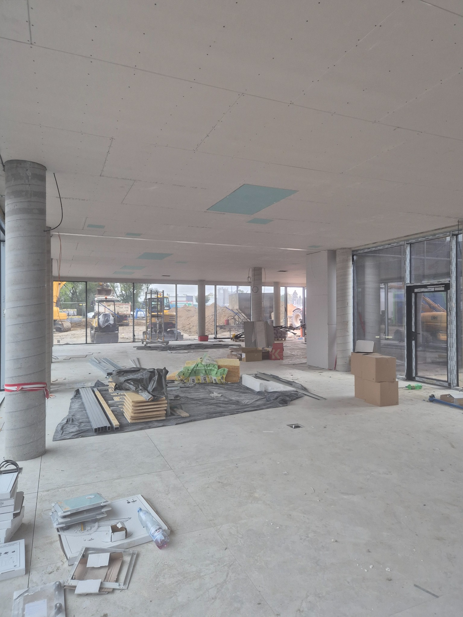 Wnętrze w trakcie prac wykończeniowych: sufit z płyt kartonowo-gipsowych, widoczne materiały budowlane i narzędzia. Przez okna widać sprzęt budowlany na zewnątrz.