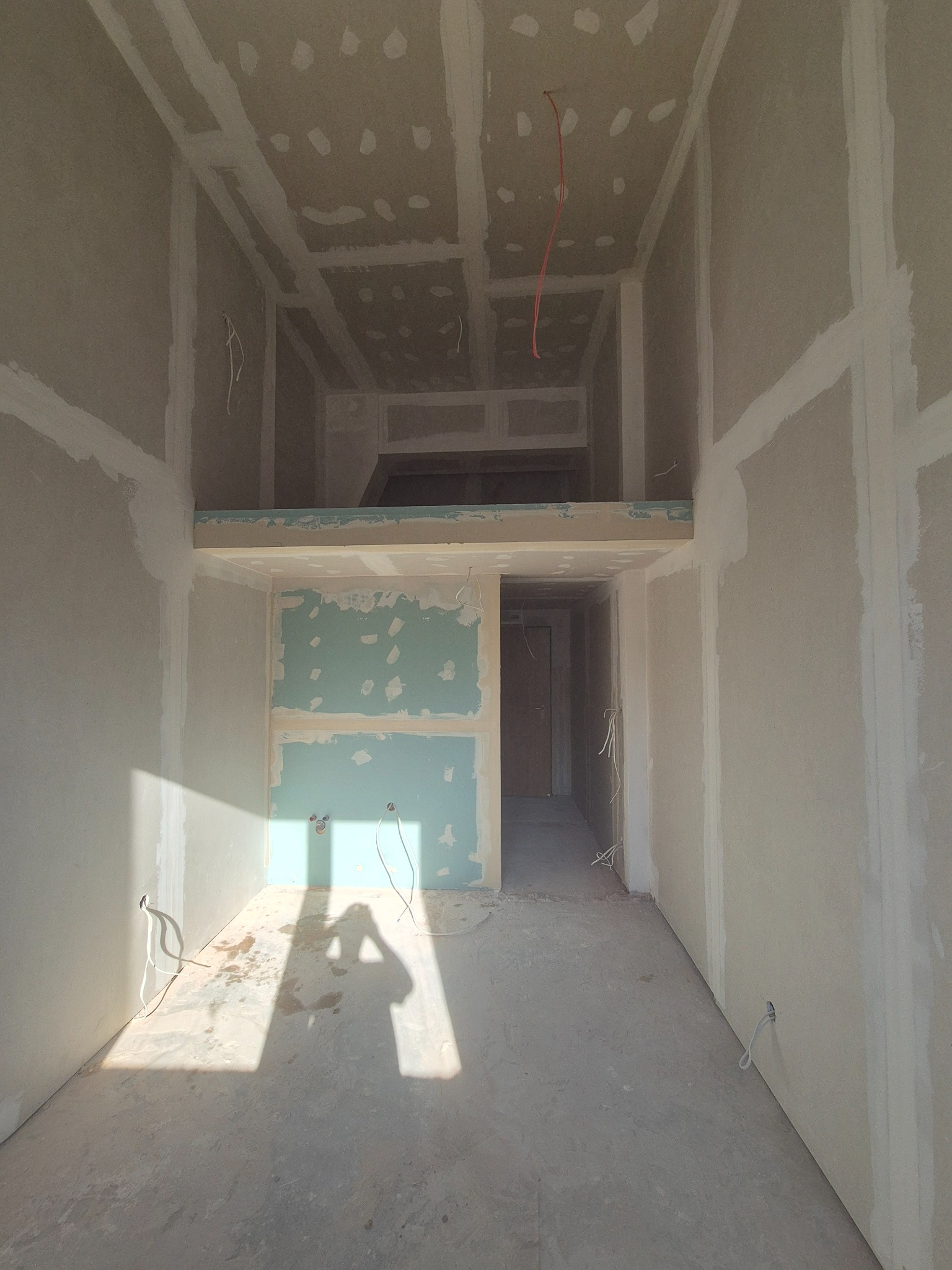 Wnętrze w trakcie prac wykończeniowych: ściany i sufit pokryte płytami gipsowo-kartonowymi, widoczne kable elektryczne, cień osoby fotografującej na podłodze.