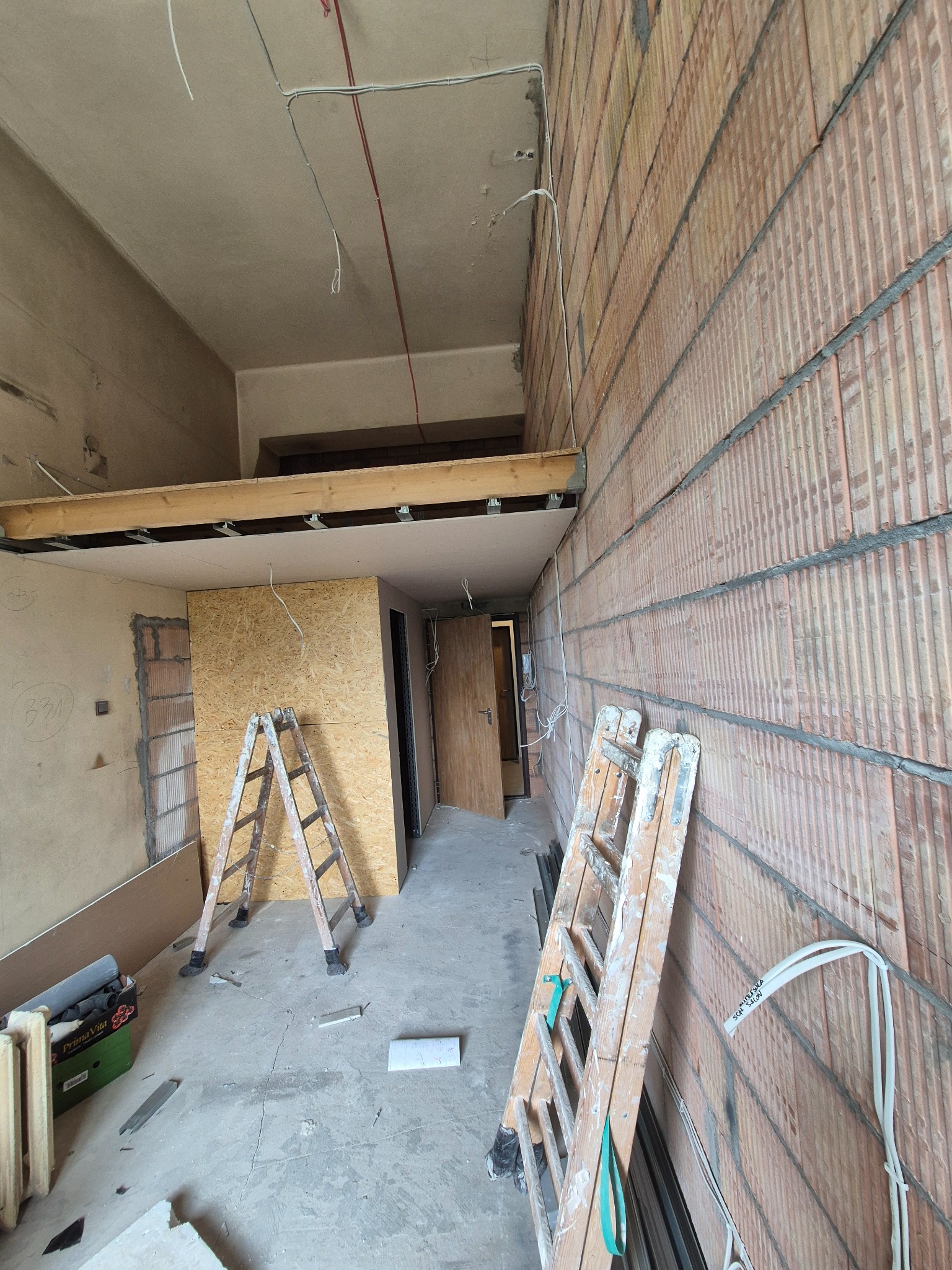 Wnętrze w trakcie prac: surowe ściany z cegły, drewniana konstrukcja sufitu, drabiny malarskie, płyta OSB. Widoczne przewody elektryczne i elementy instalacji.